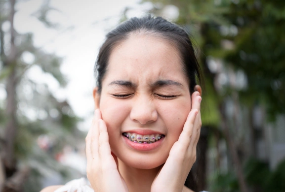 Young woman with braces wincing and holding her cheeks in discomfort outdoors