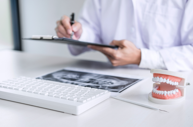 Dentist writing on a clipboard at a desk with a dental x-ray, keyboard, and teeth model