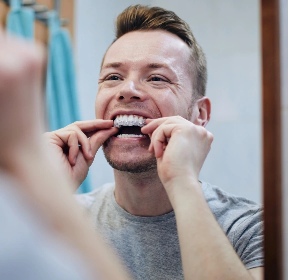 Man placing a clear aligner tray in his mouth while looking in a bathroom mirror