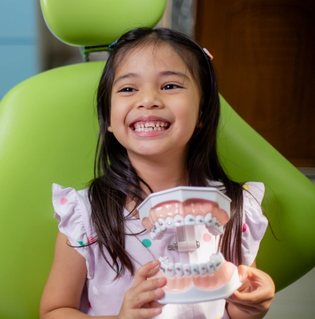 Young girl in a dental chair smiling and holding a model of teeth with braces